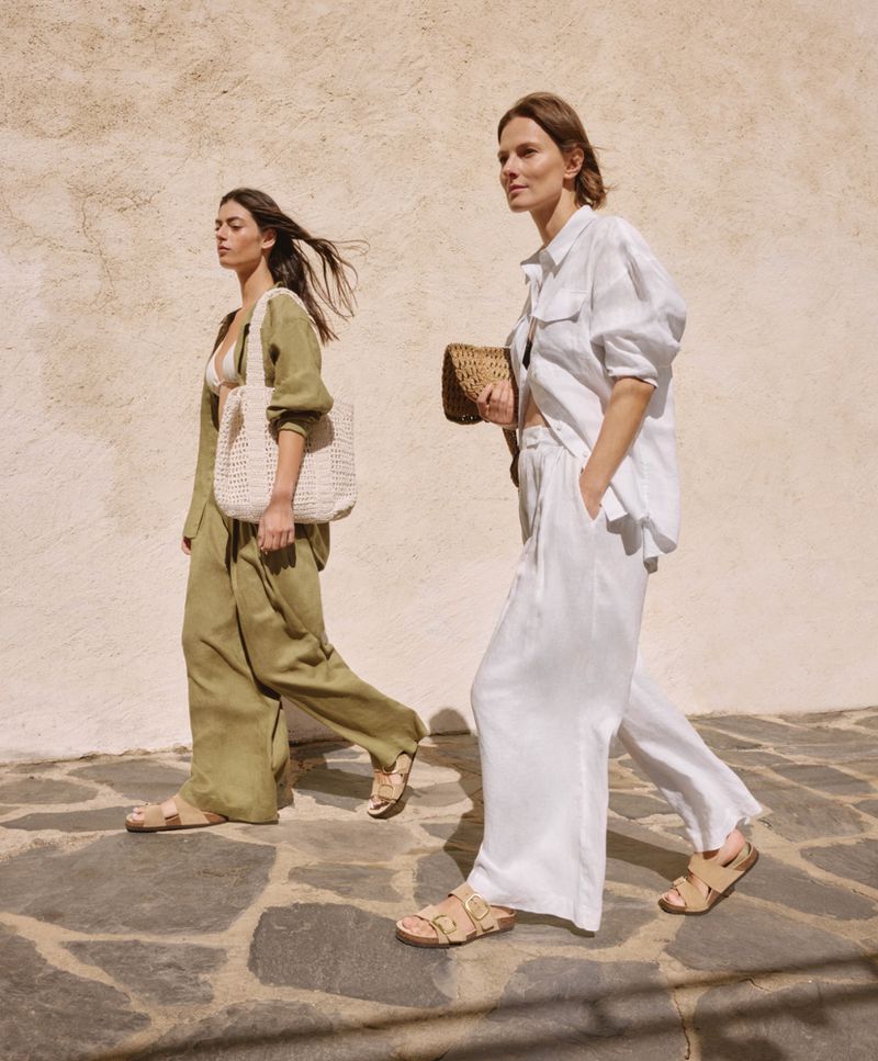 Two women in linen resort wear walk on a stone path in sunlight, one carries a woven bag, promoting fashion and relaxed summer vibes.