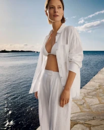 Model Tanya Ruban in an open white linen shirt, bikini top, and matching trousers standing along a stone seawall by the ocean.