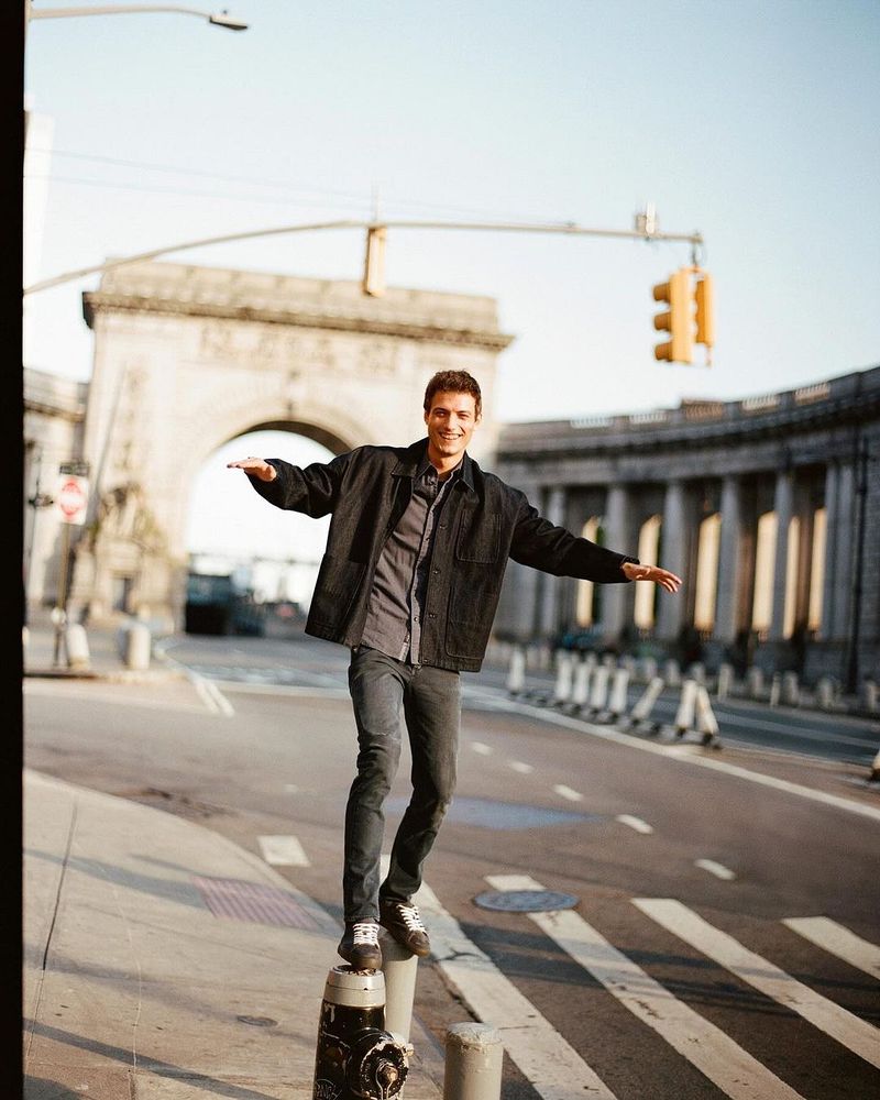 Young man balances on a post in an urban setting, wearing a jacket and jeans, with a bridge and traffic light in the background.