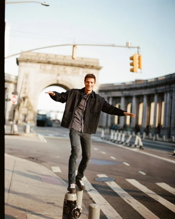 Young man balances on a post in an urban setting, wearing a jacket and jeans, with a bridge and traffic light in the background.