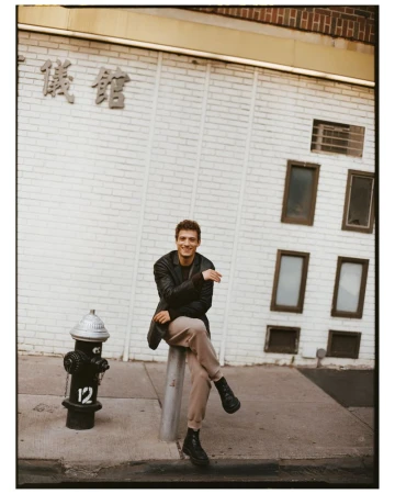 A young man sits on a metal bollard on a city sidewalk in front of a brick building with Chinese characters. He is wearing a jacket and boots.