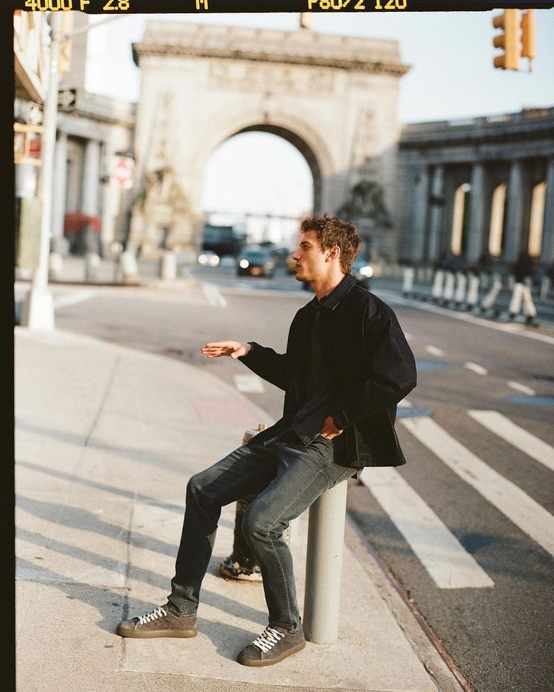 A man sits on a post in an urban setting with a monumental archway in the background. He wears dark clothing and sneakers.
