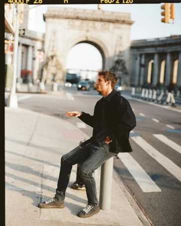 A man sits on a post in an urban setting with a monumental archway in the background. He wears dark clothing and sneakers.