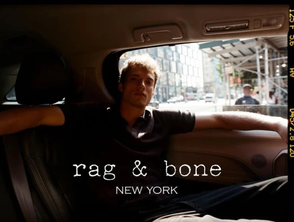 Fashion shot inside a car. A young adult male in a dark shirt sits in the back seat with a city street visible through the window.