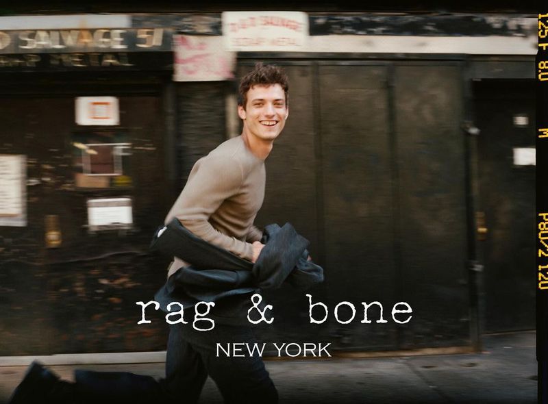 A male model in motion outdoors, smiles while holding a jacket, captured in an urban setting, featuring the 'Rag & Bone' logo.