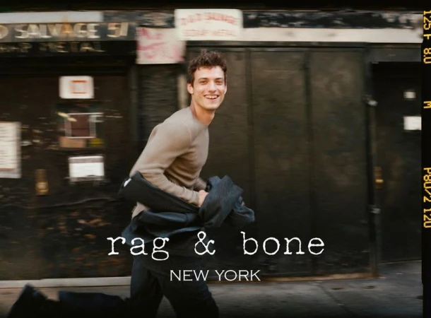 A male model in motion outdoors, smiles while holding a jacket, captured in an urban setting, featuring the 'Rag & Bone' logo.