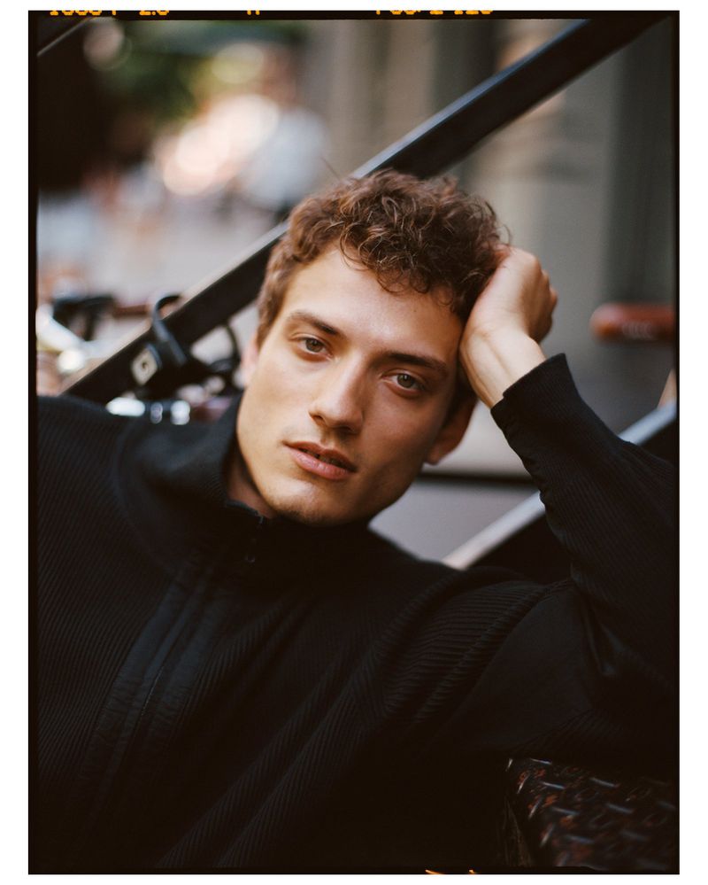 Close-up portrait of a young adult man with curly hair in a black sweater, gazing at the camera while seated on a staircase outdoors.