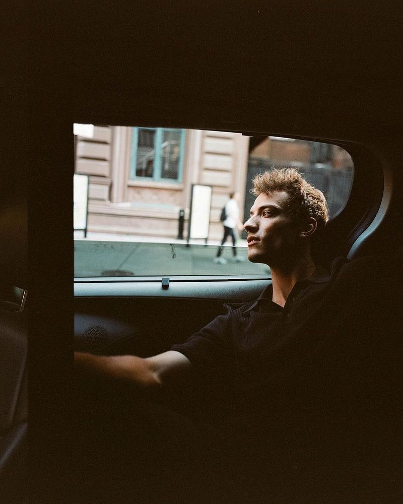 A young adult man with curly hair in a black collared shirt sits in a car with city buildings visible through the window in the background.