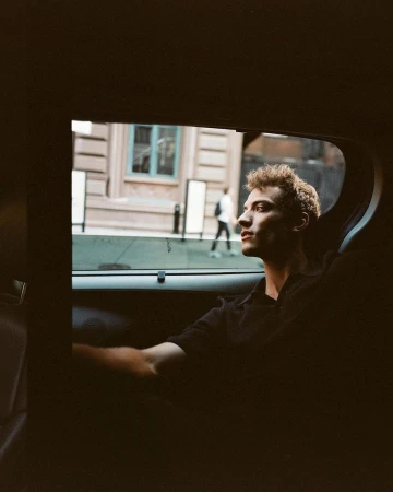 A young adult man with curly hair in a black collared shirt sits in a car with city buildings visible through the window in the background.