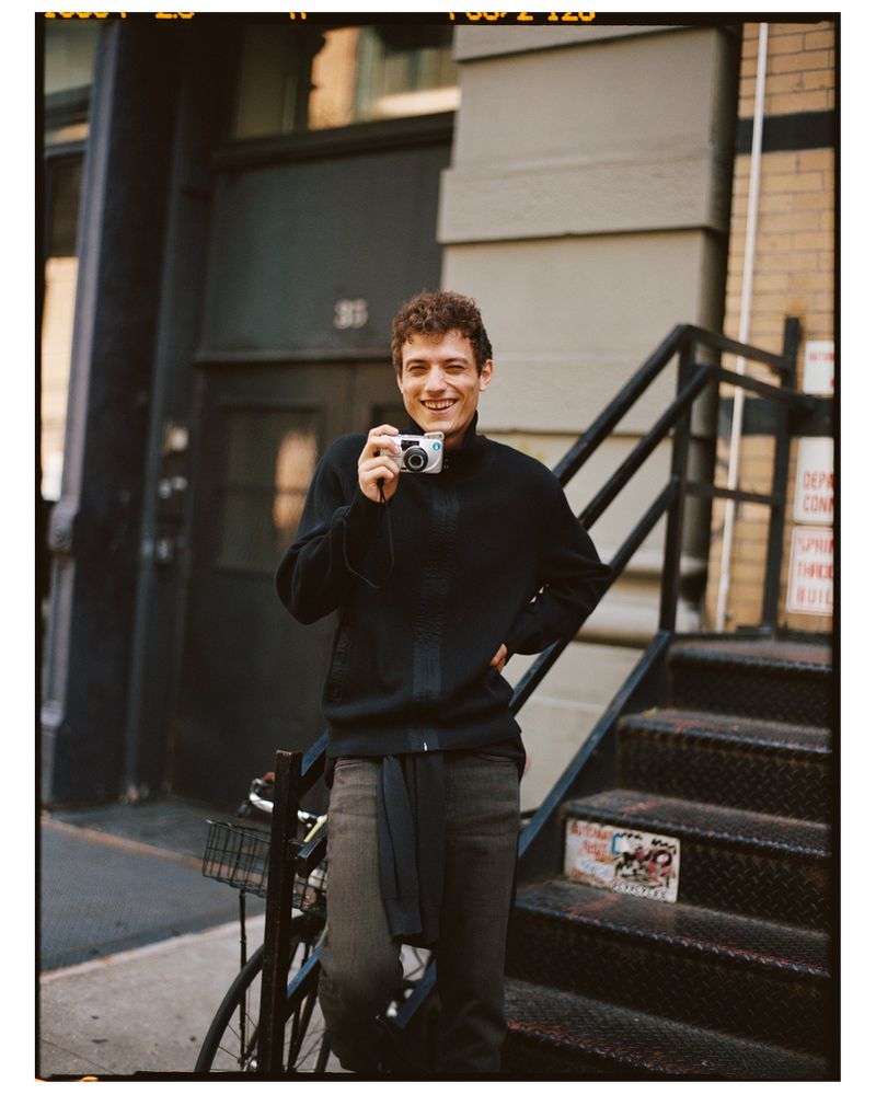 A young man with curly hair is standing on metal stairs outside a brick building, smiling and holding a compact camera.