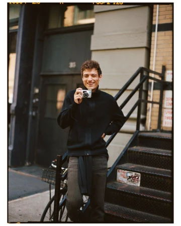 A young man with curly hair is standing on metal stairs outside a brick building, smiling and holding a compact camera.