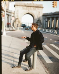 Model Serge Rigvava wearing dark jacket and jeans seated on a street bollard near an architectural archway.