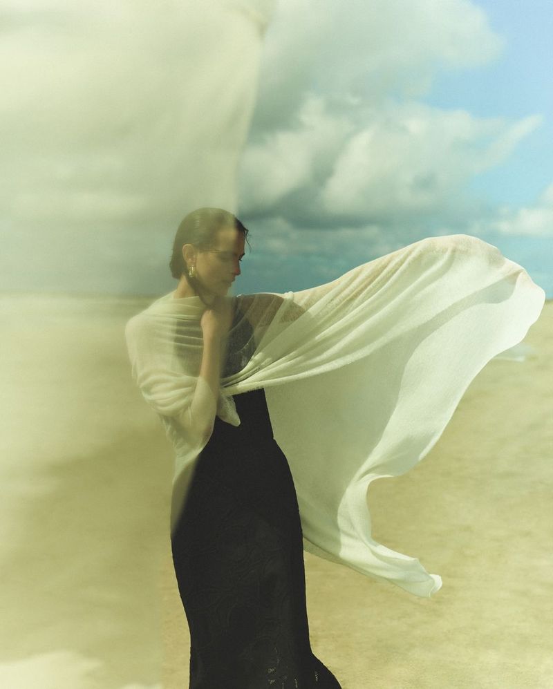 A woman in a dark dress and white wrap stands outdoors against a sky with light clouds, embodying elegance and natural beauty in soft light.