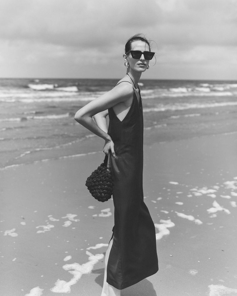 A woman in a black dress and sunglasses stands on a beach with ocean waves in the background, holding a black woven bag.