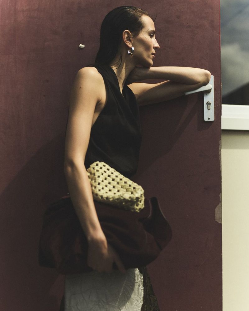 A woman leans against a door, wearing a black top, cream and brown purse, with side profile and wet hair look, standing indoors.