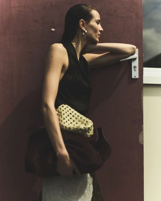A woman leans against a door, wearing a black top, cream and brown purse, with side profile and wet hair look, standing indoors.