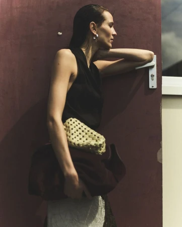 A woman leans against a door, wearing a black top, cream and brown purse, with side profile and wet hair look, standing indoors.