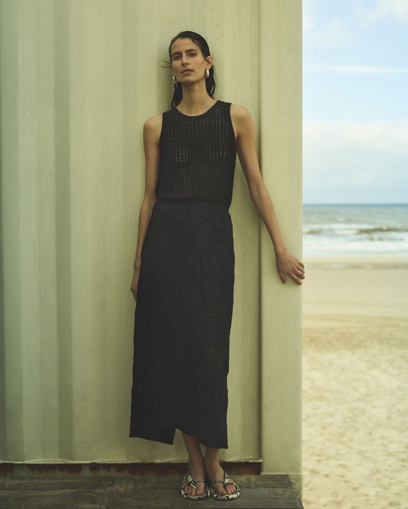 Woman in a black sleeveless knit top and long black skirt standing against a pale wall next to a sandy beach.