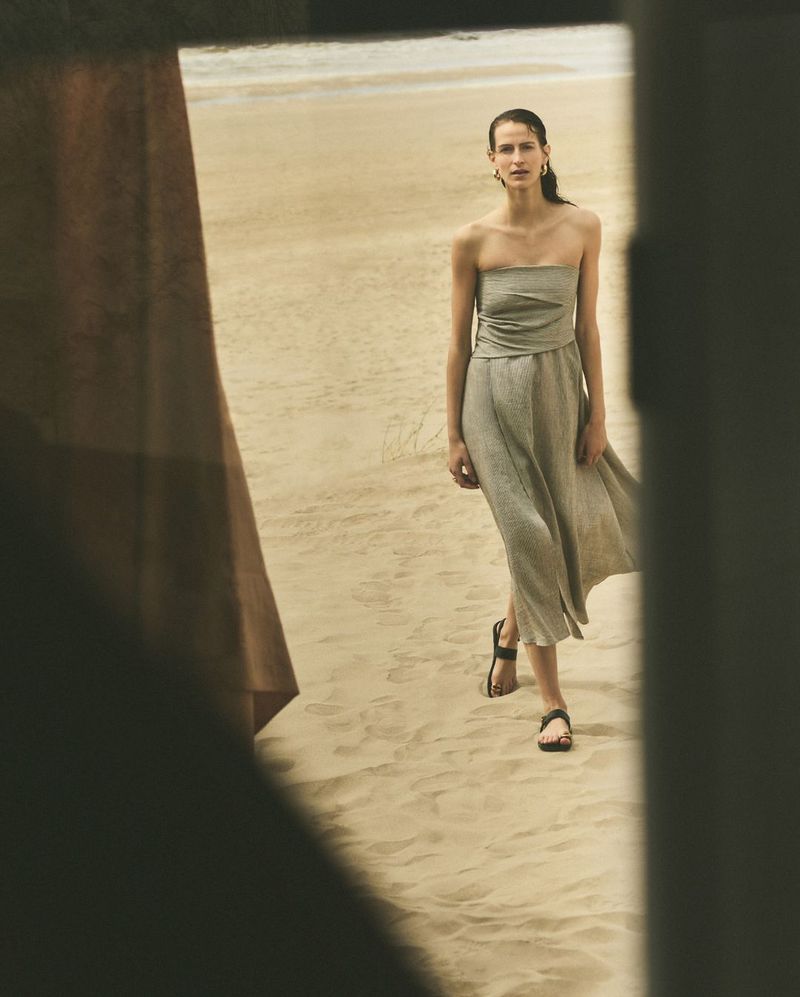 Woman in a dress walks on a sandy beach, framed by a shadowed doorway. The soft, neutral tones create a serene and stylish fashion scene.