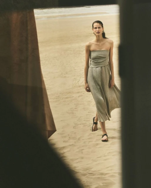Woman in a dress walks on a sandy beach, framed by a shadowed doorway. The soft, neutral tones create a serene and stylish fashion scene.