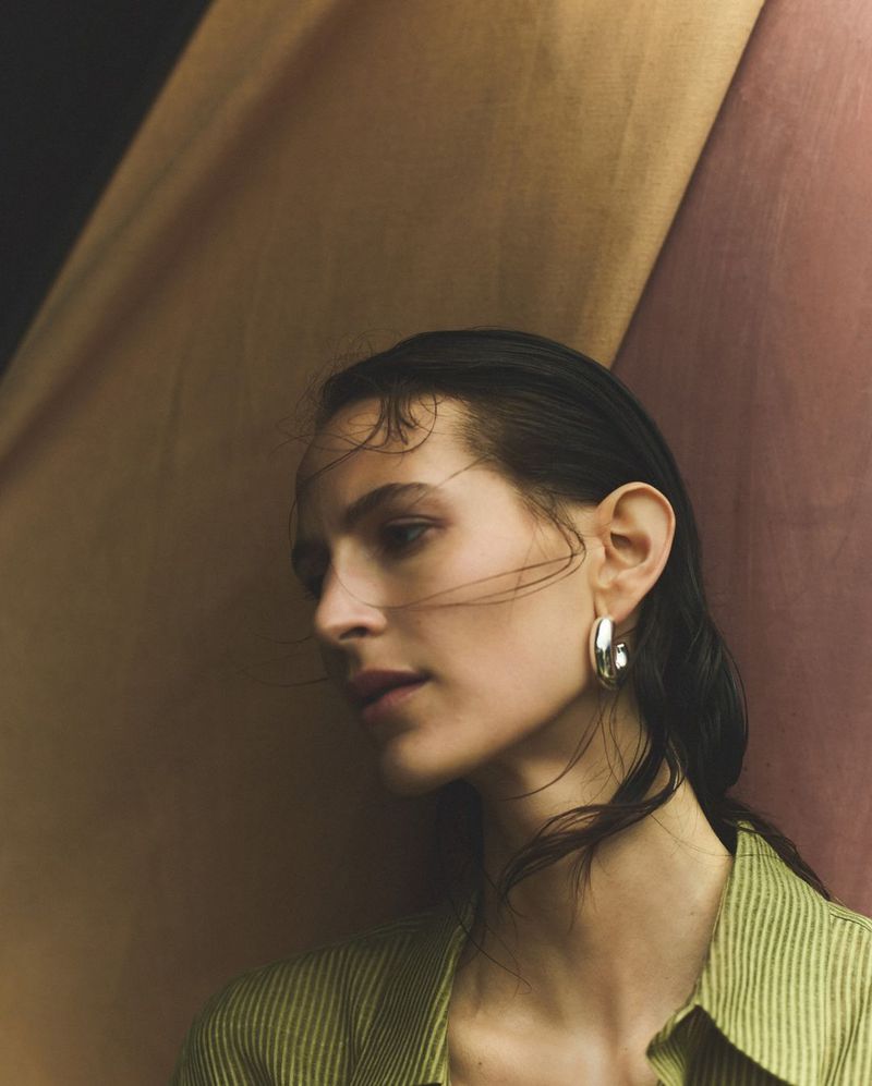 Portrait of a young woman with wet hair, wearing silver hoop earrings and a striped green shirt against a multicolored draped fabric backdrop.