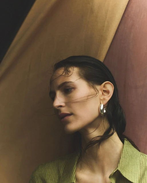 Portrait of a young woman with wet hair, wearing silver hoop earrings and a striped green shirt against a multicolored draped fabric backdrop.