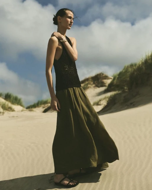 A woman stands on a sandy beach with dunes in the background, wearing a black halter top and a flowing olive green skirt.