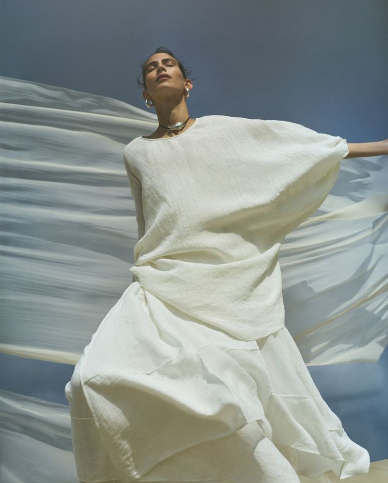 A woman in a flowing white dress stands against a sky and fabric backdrop, embodying a light, airy fashion aesthetic.
