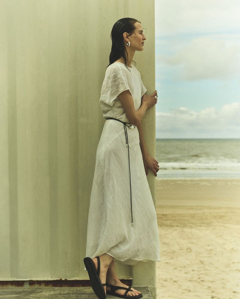 Woman in white linen dress stands by architectural wall on a beach with ocean view. Neutral fashion shot in natural daylight.