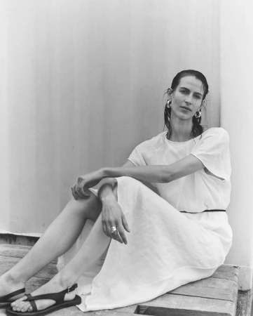 A woman in a flowing white short-sleeve dress and black flat sandals sits on a wooden surface against a neutral wall.