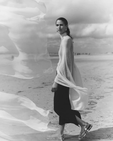 Model walking on a sandy beach wearing a sheer high-neck top, dark skirt, and flat sandals beside billowing translucent fabric.