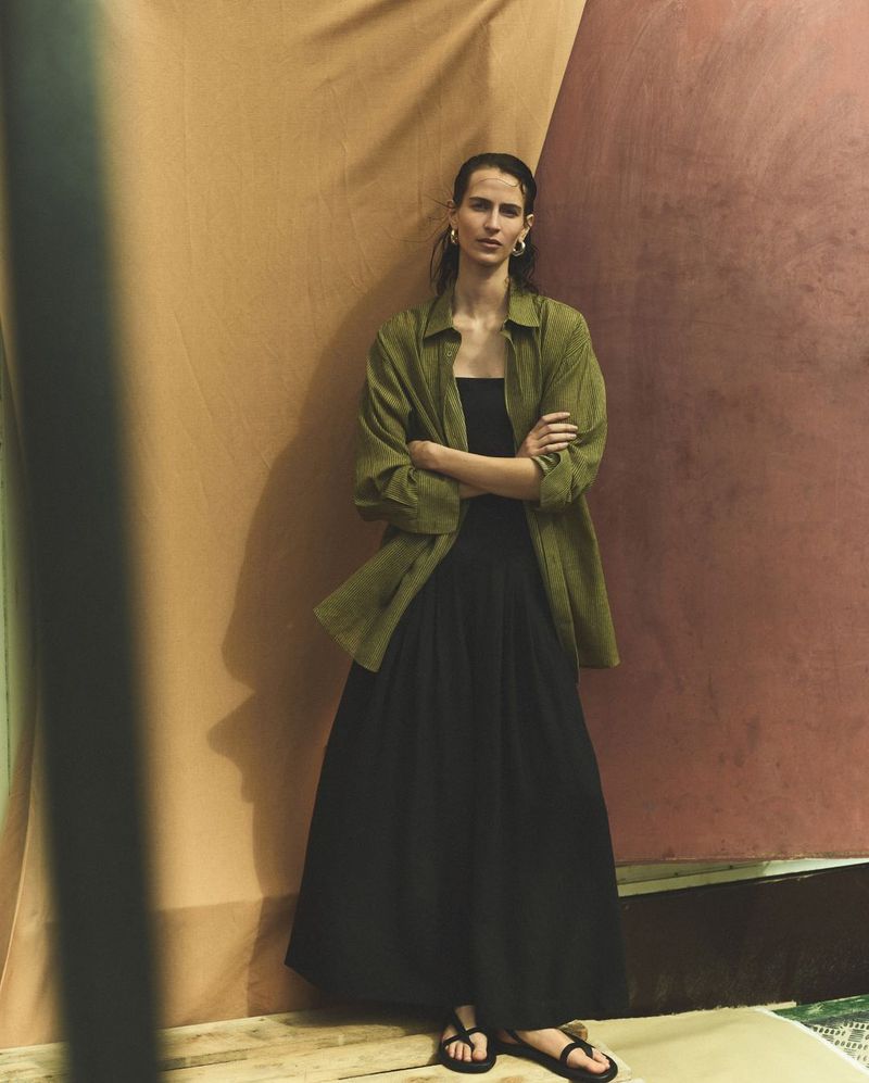 Fashionable woman in green shirt and black dress poses in front of a neutral backdrop, looking confidently at the camera.