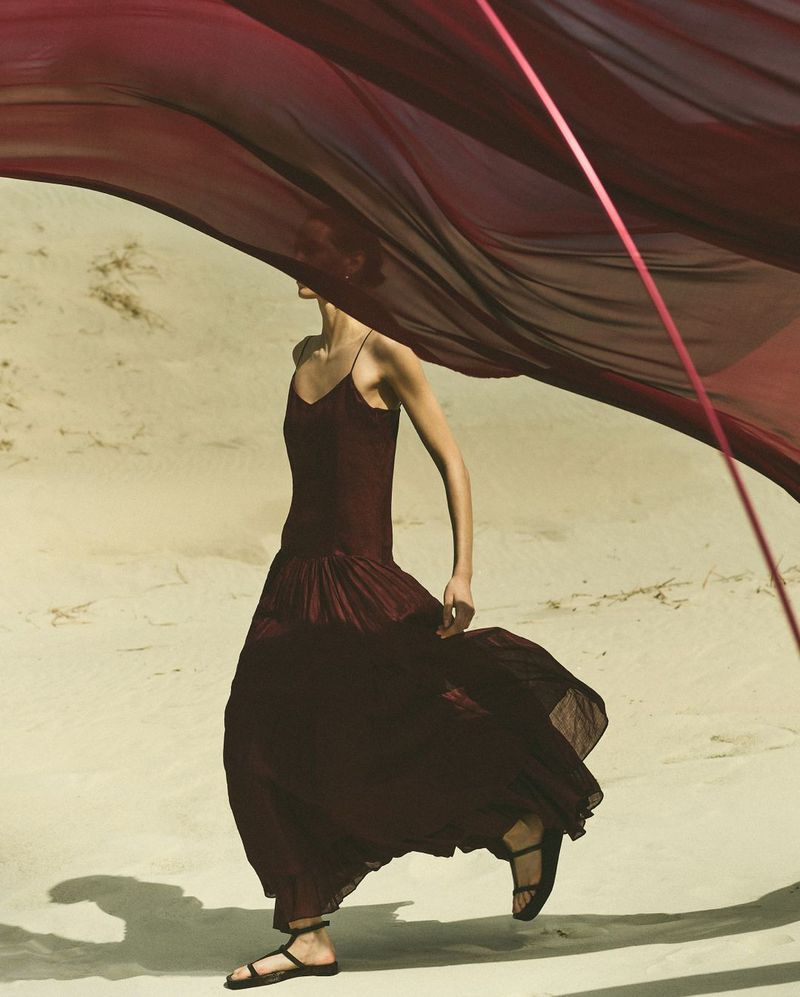 An adult female fashion model in a burgundy dress walks on sand, a large burgundy fabric flowing in the wind, outdoor, lifestyle photoshoot.