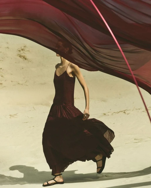 An adult female fashion model in a burgundy dress walks on sand, a large burgundy fabric flowing in the wind, outdoor, lifestyle photoshoot.