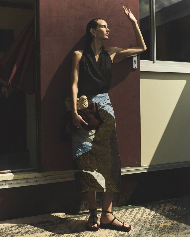 Elegant adult woman in a black top and patchwork skirt stands outside a beige trailer, shielding her eyes from sunlight in a fashion shot.