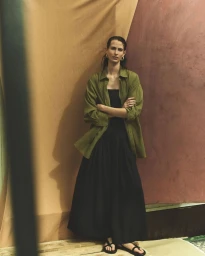 Model wearing an open olive green shirt over a black maxi dress standing with crossed arms against a draped studio backdrop.