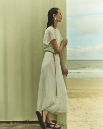 Woman in a flowing white linen dress and black flat sandals standing next to a structure overlooking a sandy beach and ocean.