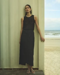 Woman in a black sleeveless knit top and long black skirt standing against a pale wall next to a sandy beach.