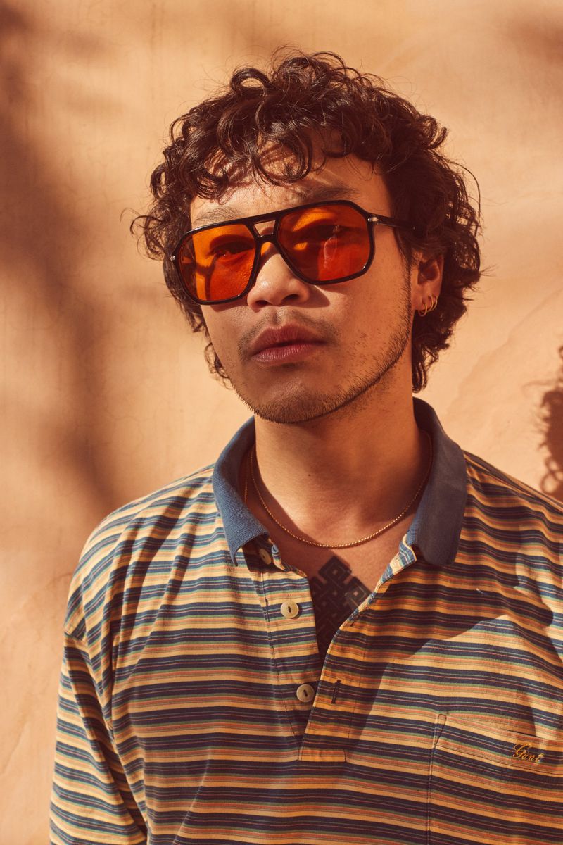 Portrait of a young adult with curly hair wearing orange sunglasses and a striped polo shirt, posing in front of a textured wall.