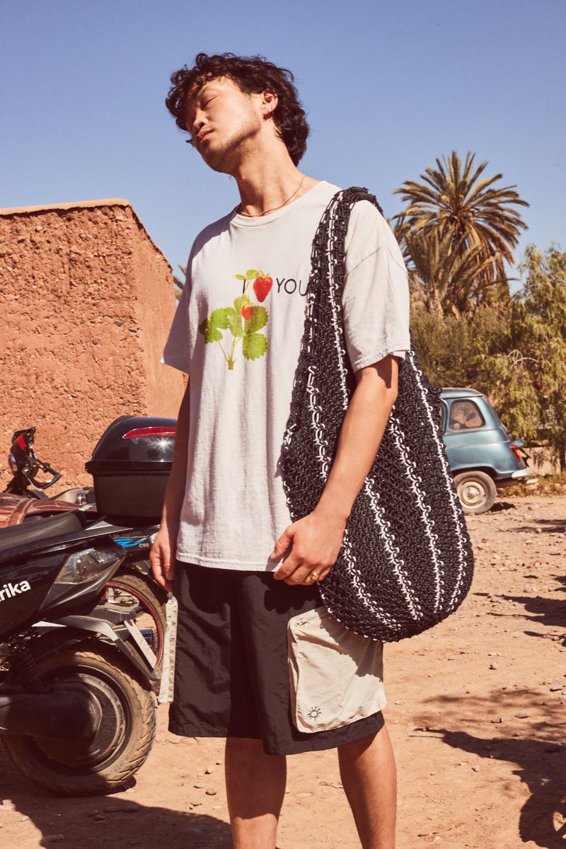 Young man in a t-shirt with a strawberry graphic and black tote bag poses outdoors in the sun. A moped and car are nearby.