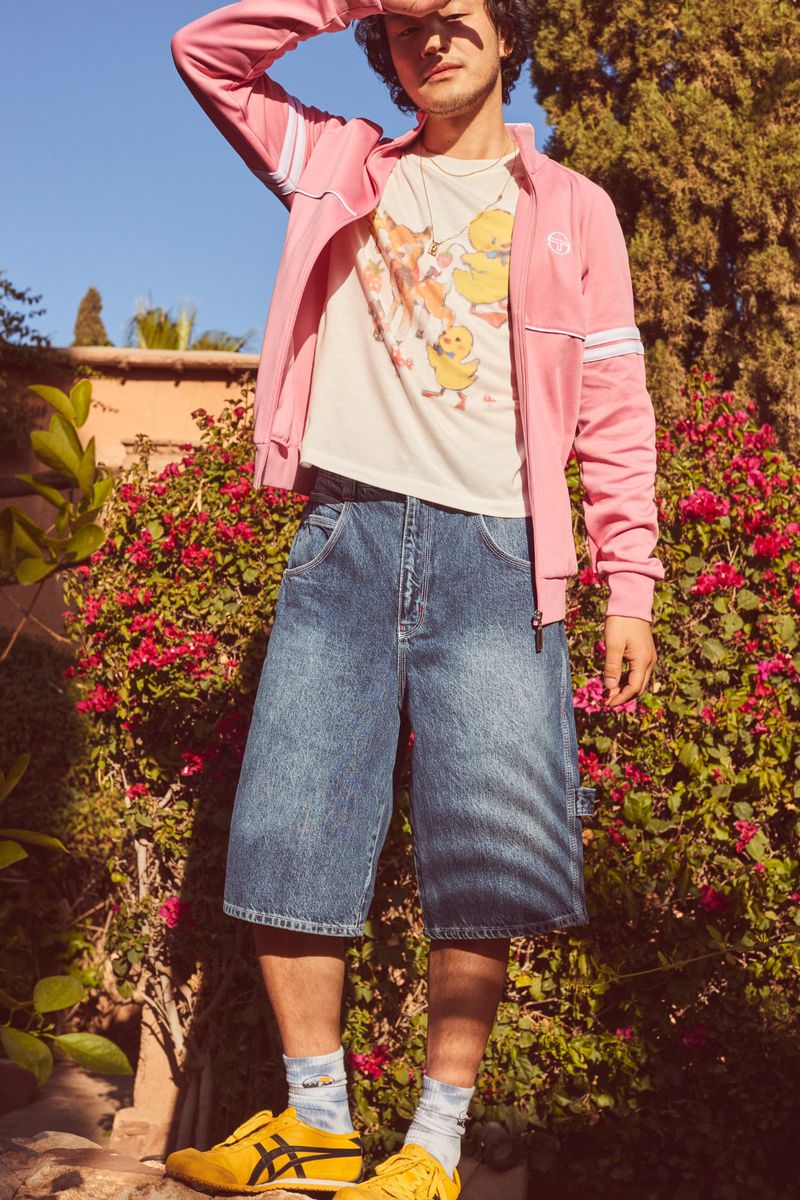 Man stands in garden wearing a pink track jacket, graphic t-shirt, wide denim shorts, and yellow sneakers in natural sunlight.