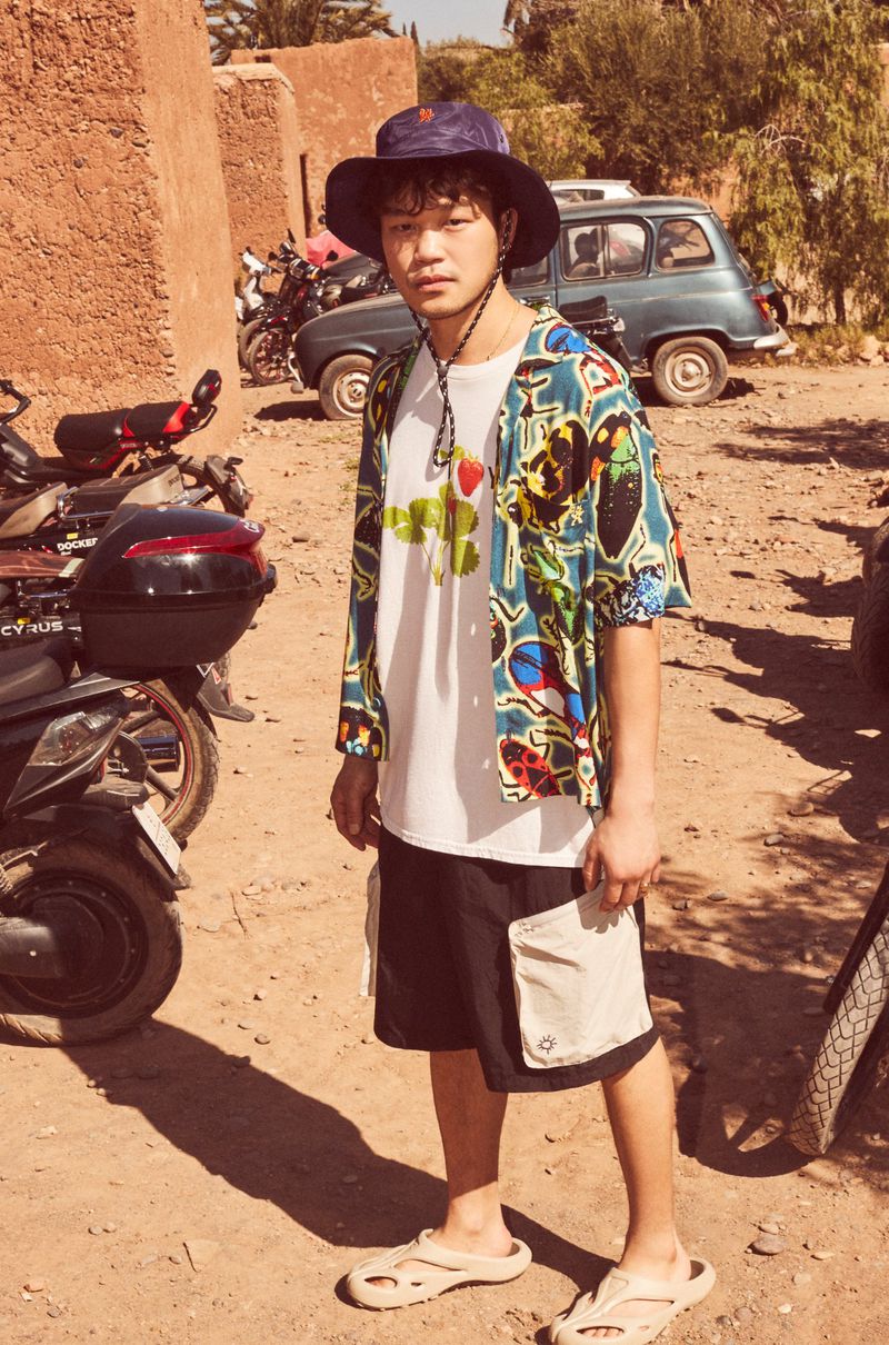 An Asian man in a hat and patterned shirt stands outdoors on a sunny day, with motorcycles and a car in the background, wearing shorts and sandals.
