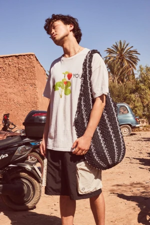 Model wearing a graphic t-shirt and dark cargo shorts carrying a large woven shoulder bag outdoors.