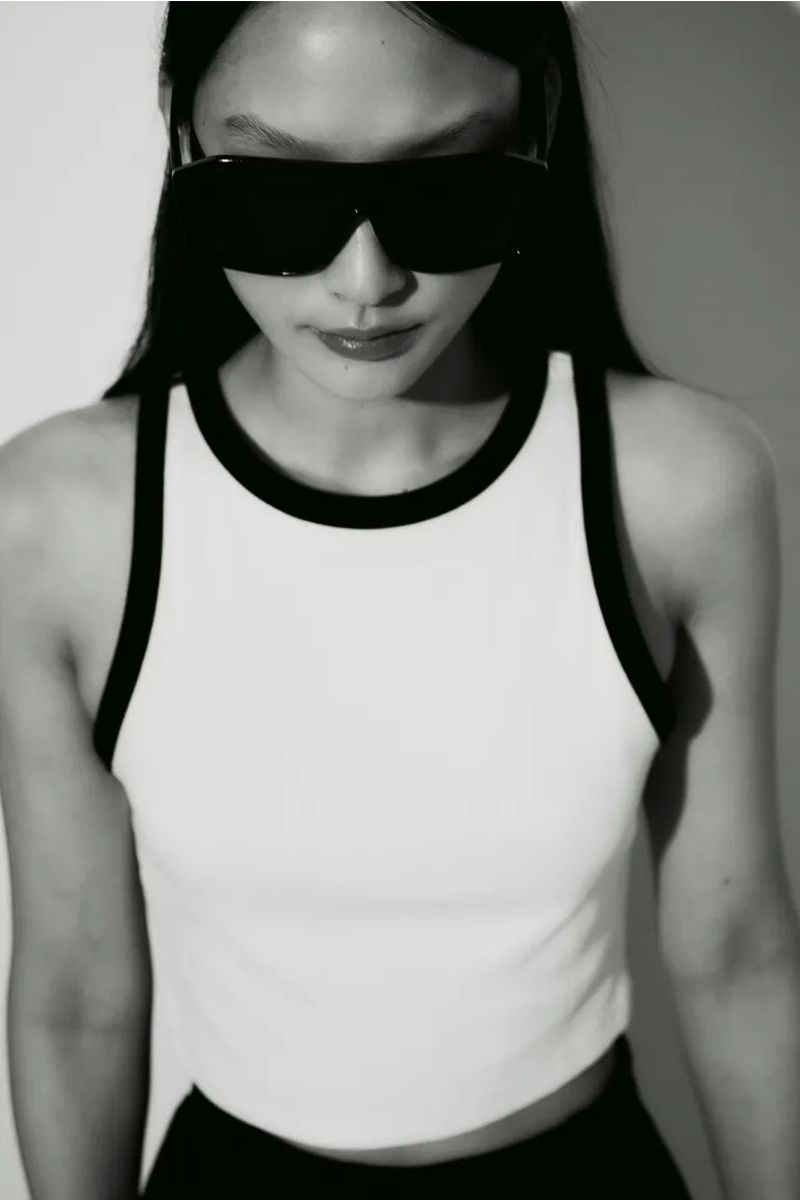 Model Yoonmi Sun wearing oversized black shield sunglasses and a white tank top with black contrast trim against a white backdrop.