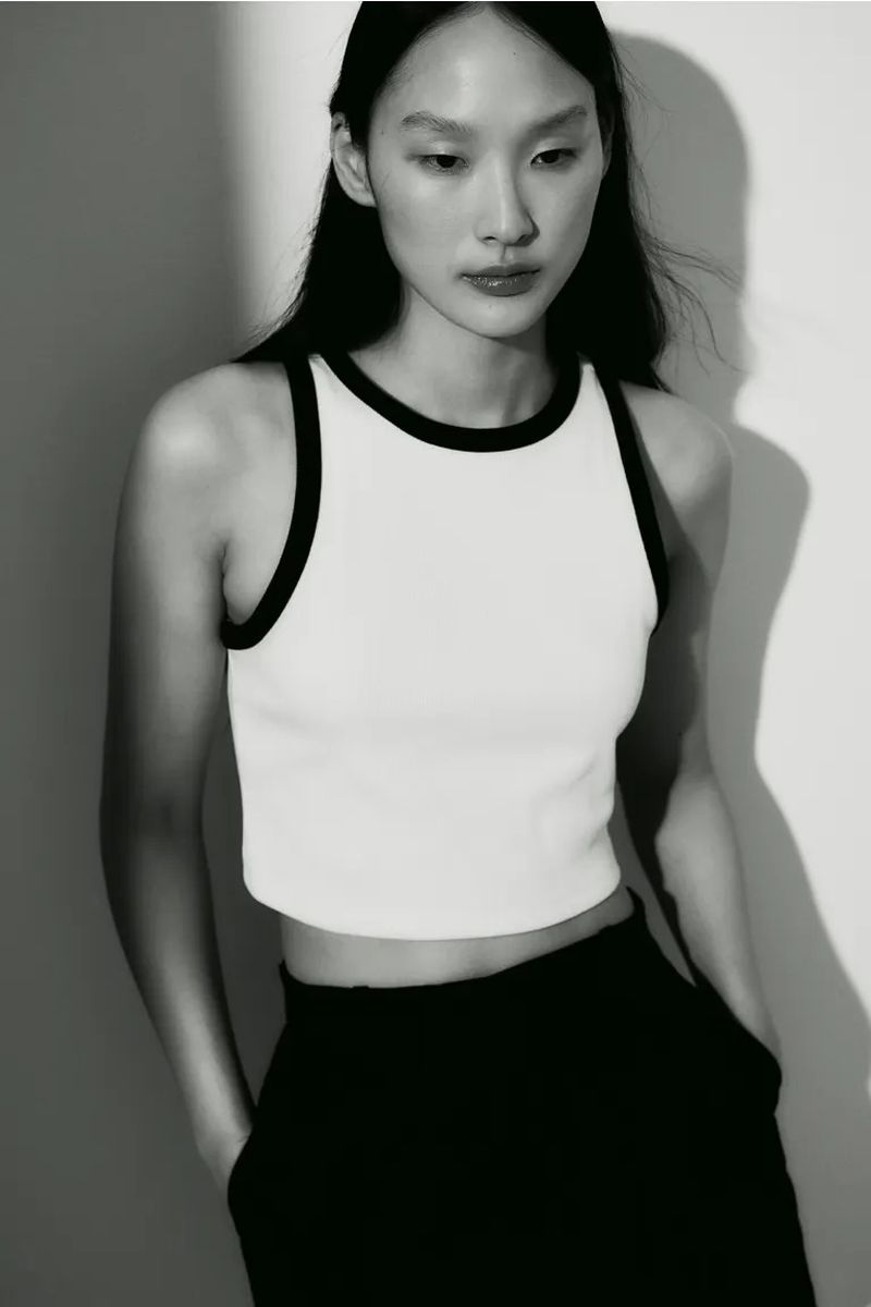 Monochrome studio portrait of an Asian woman wearing a cropped tank top and black pants, standing against a neutral wall.