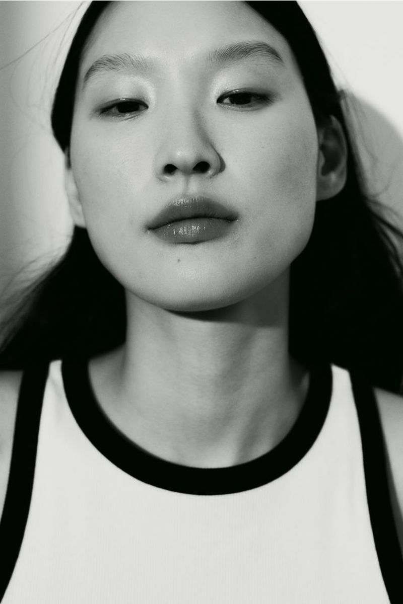 Close-up black and white portrait of a young adult Asian woman with dark hair, wearing a white top with black trim, against a white wall.