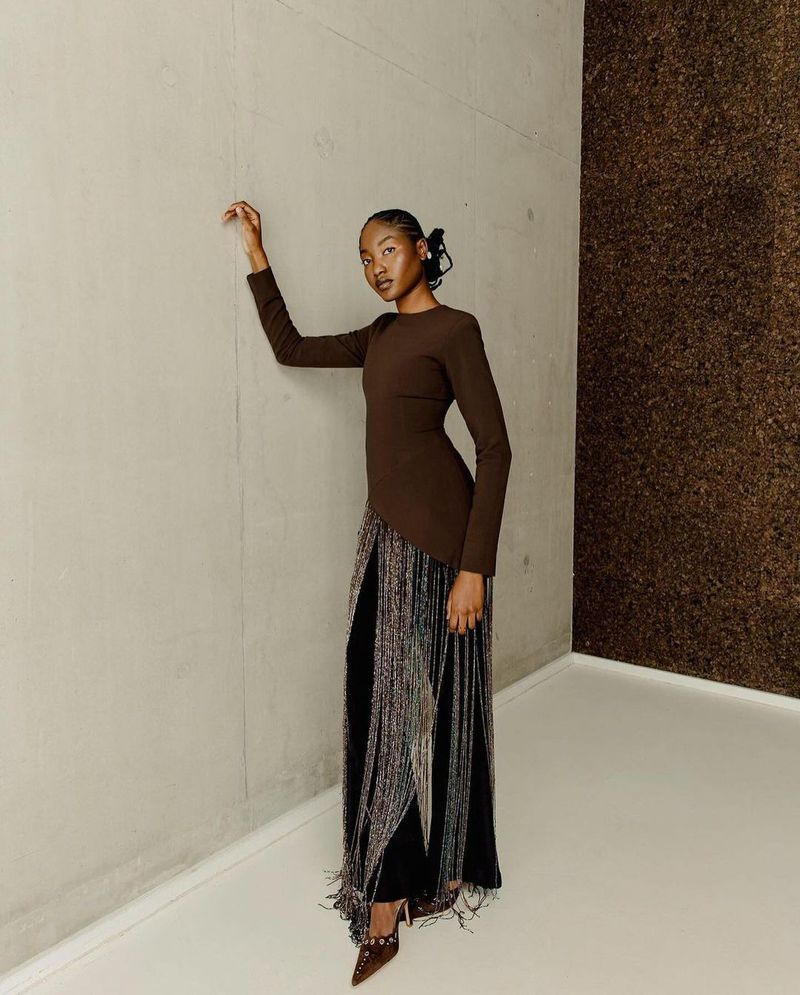 Elegant woman in a long-sleeved brown dress and fringe skirt poses against a two-toned wall inside a studio setting.