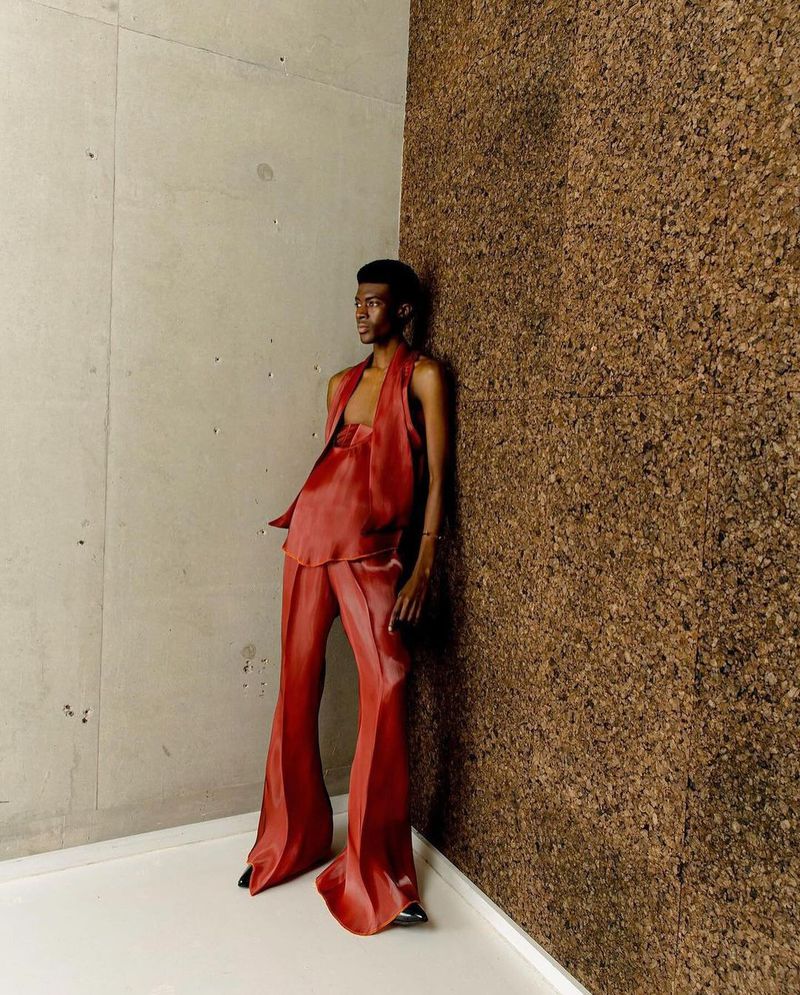 An adult male model poses in terracotta satin halter top and flared pants against a corner of textured concrete and brown cork paneling.