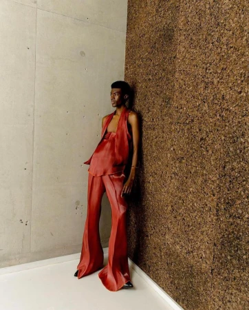 An adult male model poses in terracotta satin halter top and flared pants against a corner of textured concrete and brown cork paneling.