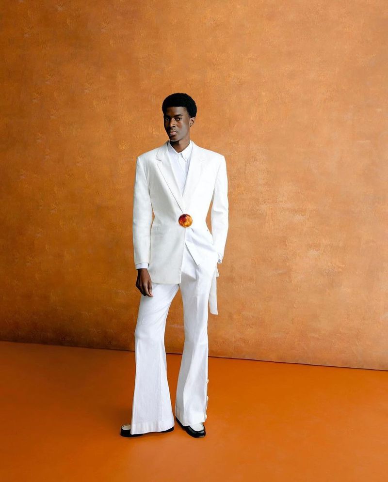 Full-length shot of a Black male model in white suit posing against orange backdrop in studio, displaying retro style and modern fashion.